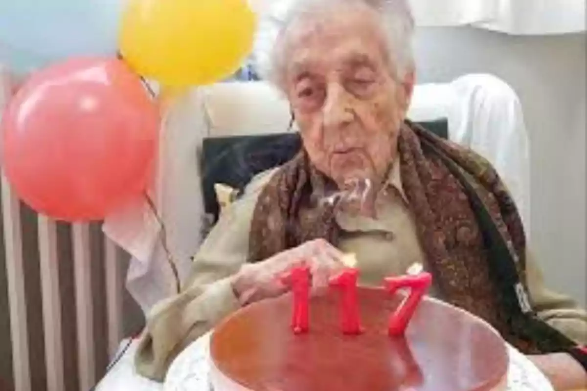 Una persona gran bufant les espelmes d'un pastís d'aniversari amb el número 117, amb globus de colors al fons.