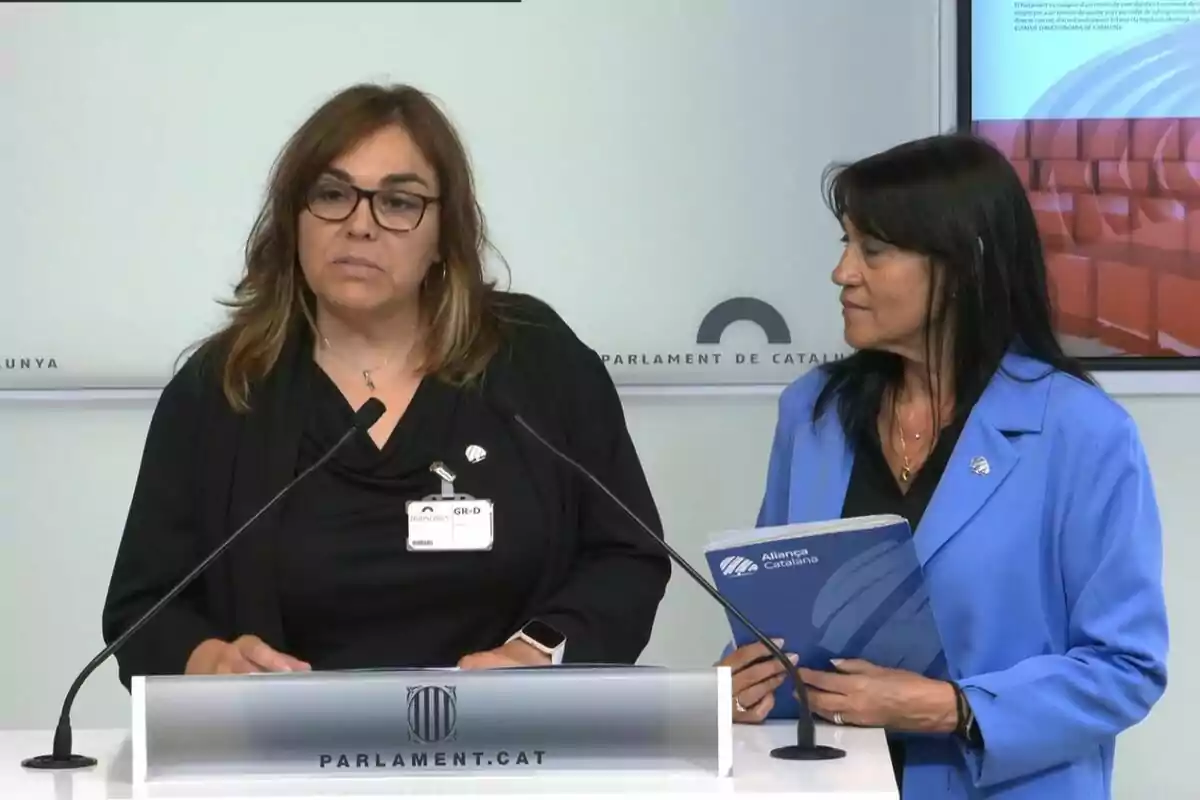 Dues dones en una conferència de premsa al Parlament de Catalunya, una d'elles parla mentre l'altra sosté un document d'Aliança Catalana. Dues dones en una conferència de premsa al Parlament de Catalunya, una d'elles parla mentre l'altra sosté un document d'Aliança Catalana.
