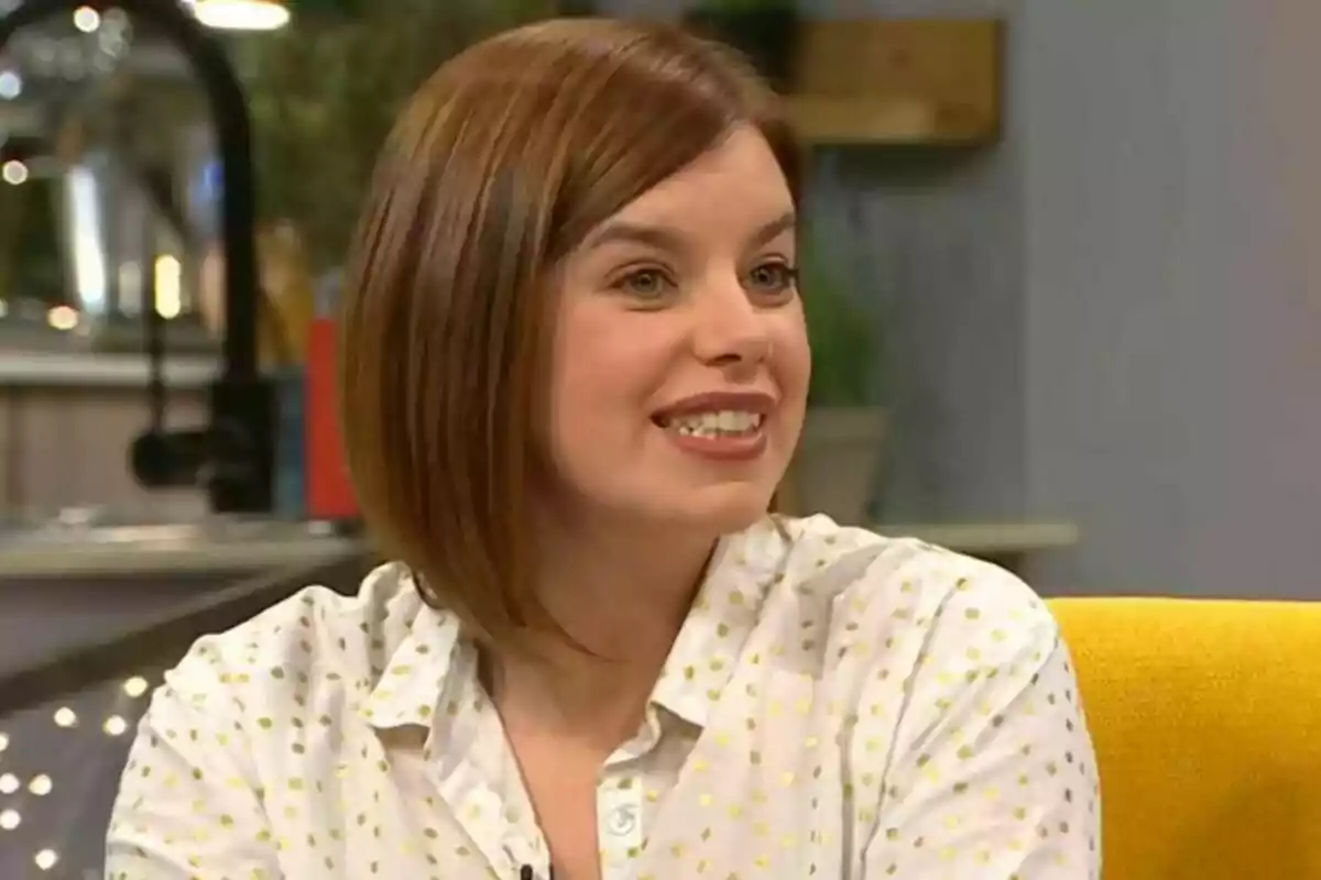 Woman with short brown hair and white shirt with yellow dots smiling as she sits on a yellow armchair in an indoor setting.