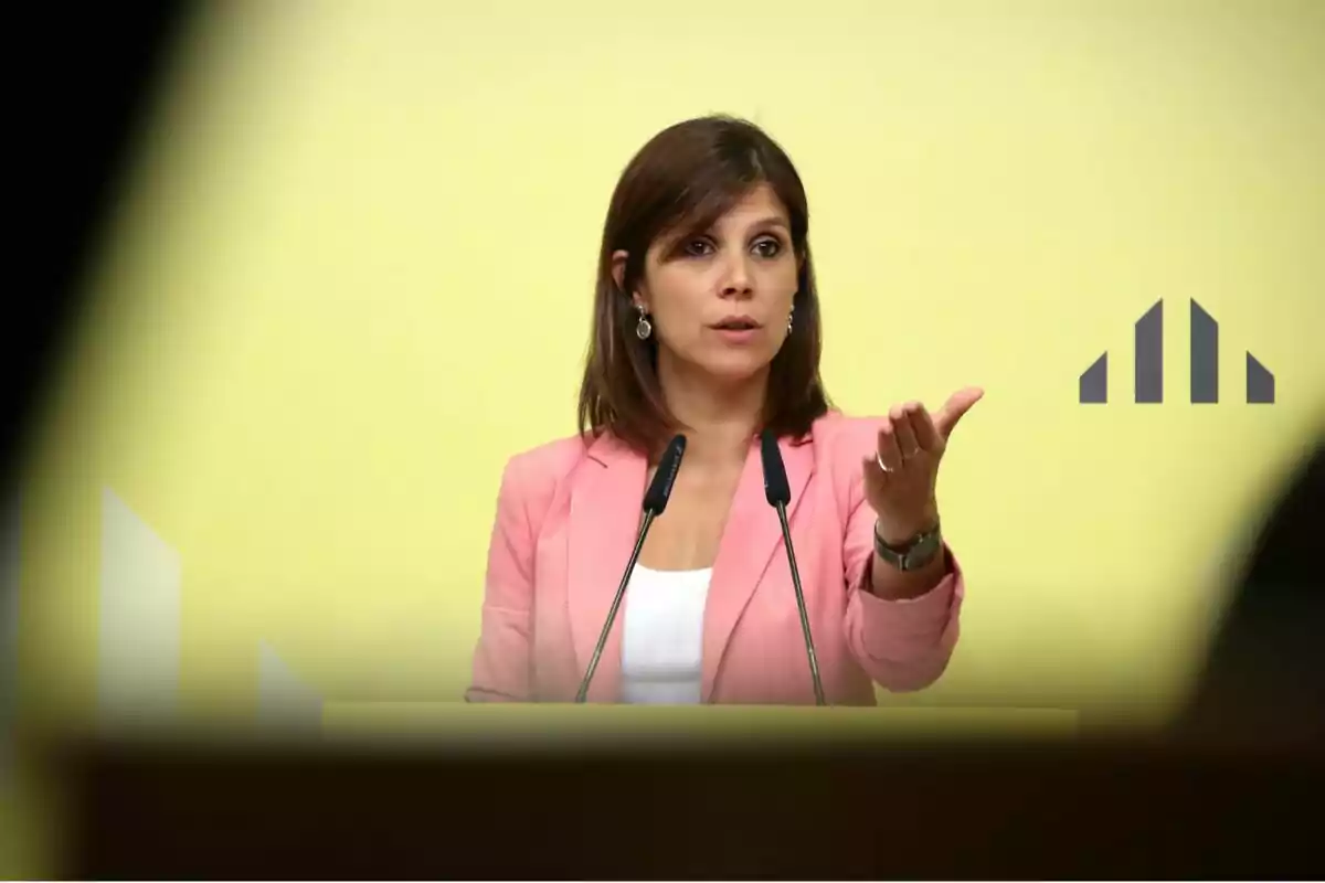 Dona amb jaqueta rosa parlant en un podi amb micròfons davant d'un fons groc.