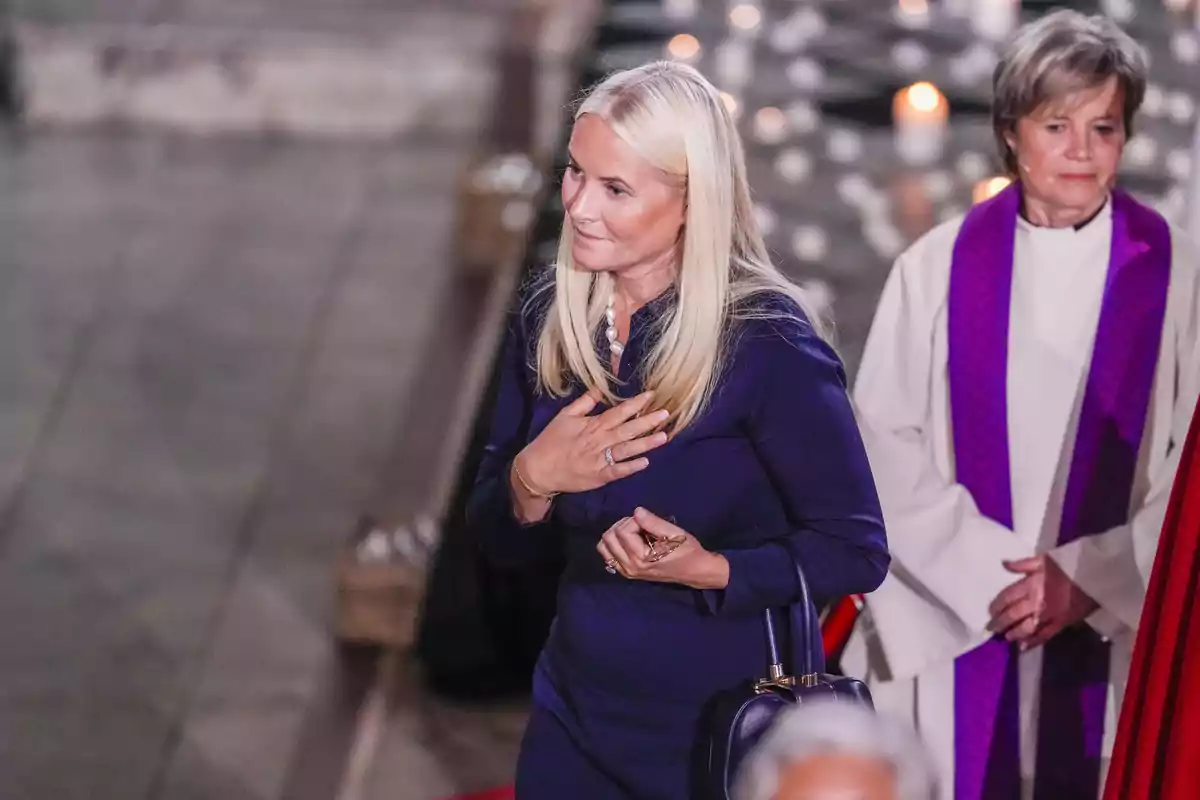 Mette-Marit con vestido azul oscuro está de pie en un entorno religioso, con una mano en el pecho y una expresión solemne, mientras otra persona con vestimenta clerical está detrás de ella. Mette-Marit con vestido azul oscuro está de pie en un entorno religioso, con una mano en el pecho y una expresión solemne, mientras otra persona con vestimenta clerical está detrás de ella.