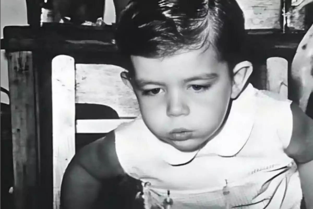 Un niño pequeño con cabello oscuro y expresión seria, vestido con una camisa blanca, en una fotografía en blanco y negro. Un niño pequeño con cabello oscuro y expresión seria, vestido con una camisa blanca, en una fotografía en blanco y negro.
