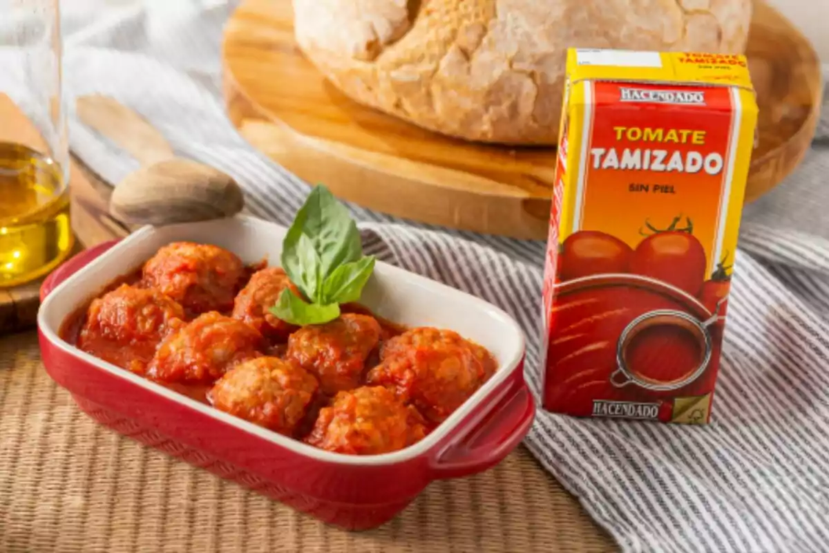 Un plat de mandonguilles amb salsa de tomàquet al costat d'un envàs de tomàquet tamisat sense pell i una fogassa de pa al fons. Un plat de mandonguilles amb salsa de tomàquet al costat d'un envàs de tomàquet tamisat sense pell i una fogassa de pa al fons.