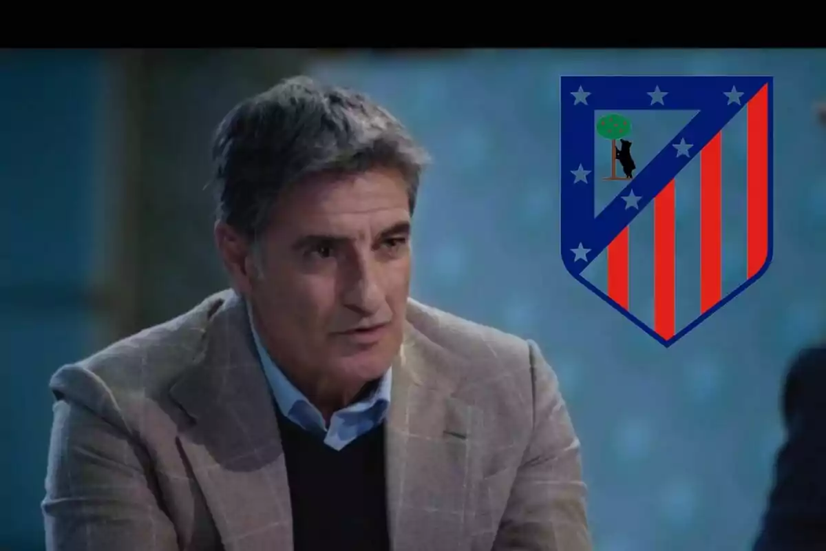 Hombre con saco gris y camisa azul junto al escudo del Atlético de Madrid.