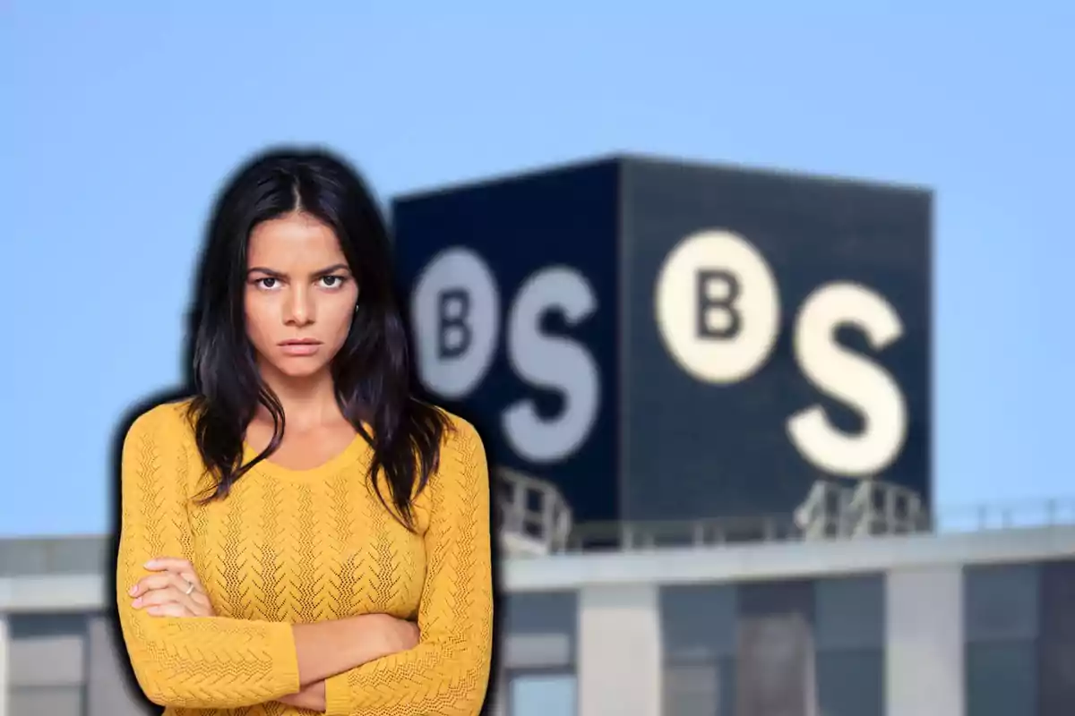 Mujer con suéter amarillo de brazos cruzados frente a un edificio con las letras B y S en el fondo.