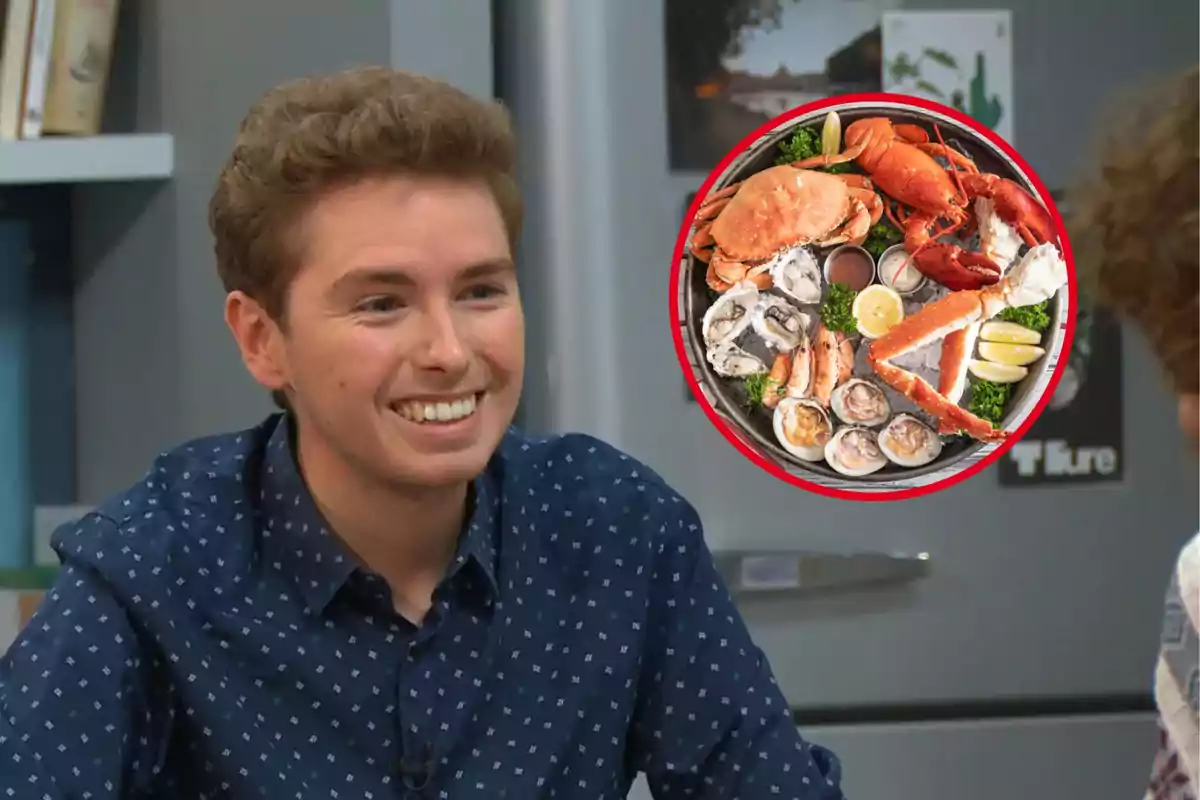 Un hombre sonriente con una camisa azul de lunares está sentado frente a una mesa y hay una imagen superpuesta de un plato de mariscos variados.