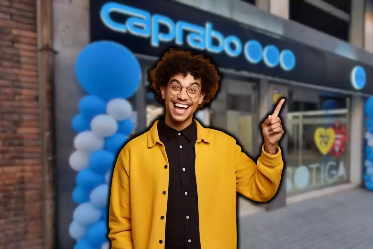 Un hombre sonriente con gafas y chaqueta amarilla señala hacia un letrero de una tienda con decoración de globos azules y blancos.