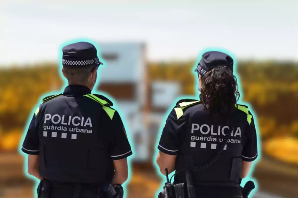 Dos agents de la policia urbana d?esquena, amb uniformes foscos i detalls reflectants, observen un vehicle borrós al fons en un entorn al?aire lliure.