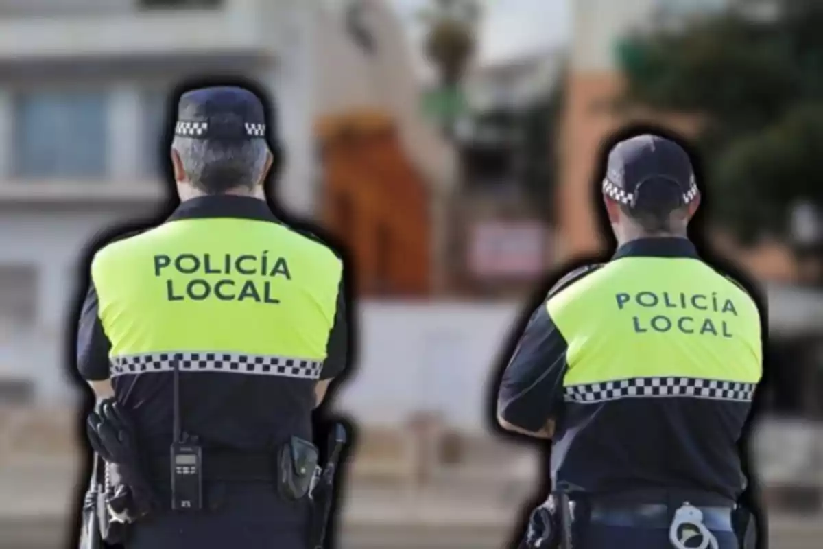 Dos agentes de policía local de espaldas con chalecos reflectantes amarillos en un entorno urbano.