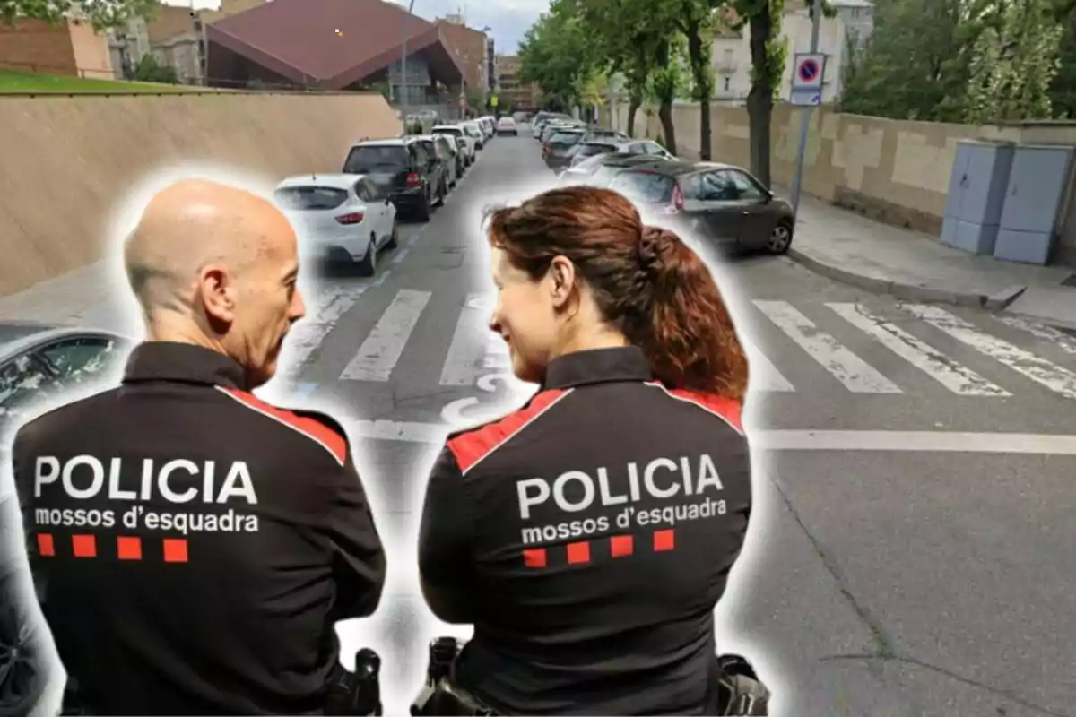 Dos agentes de la policía de los Mossos d'Esquadra de espaldas en una calle con coches estacionados y un paso de peatones.