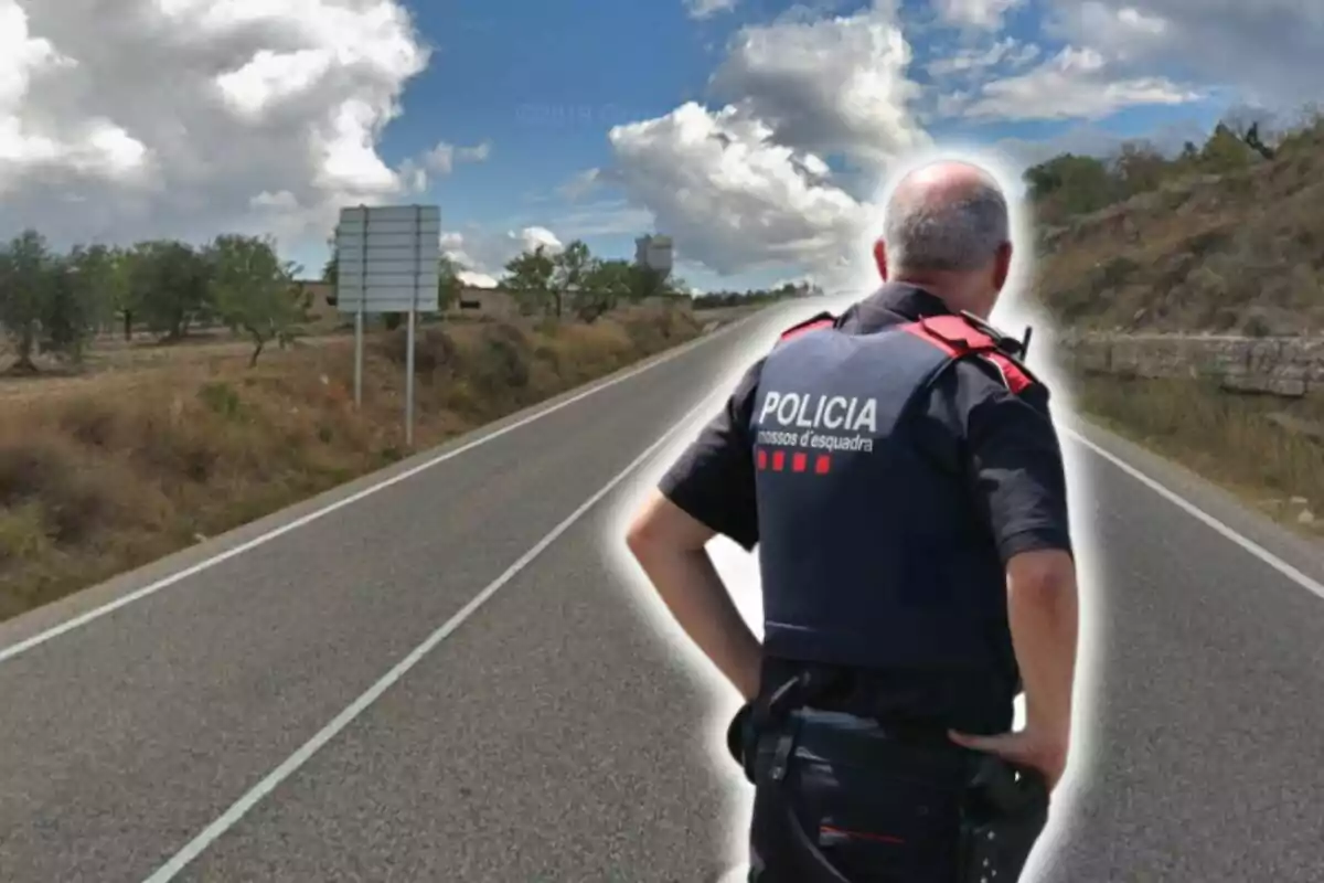 Un policia dels Mossos d'Esquadra d'esquena a una carretera rural sota un cel parcialment ennuvolat.