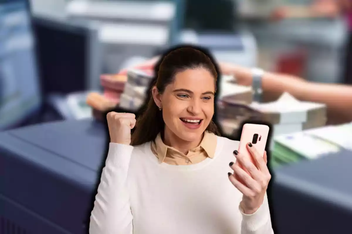 Mujer sonriendo y levantando el puño mientras mira su teléfono móvil, con un fondo de oficina y pilas de papeles.