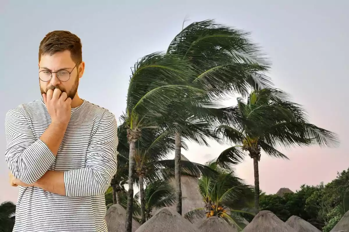 Hombre pensativo con gafas y camiseta de rayas frente a palmeras movidas por el viento.