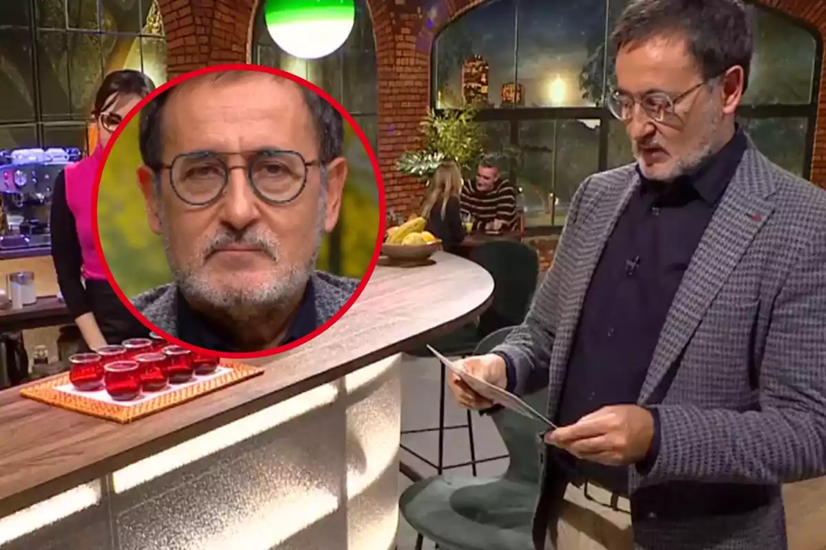 Un home amb ulleres i barba llegeix un paper en un estudi de televisió amb una taula de begudes vermelles i una dona al fons.