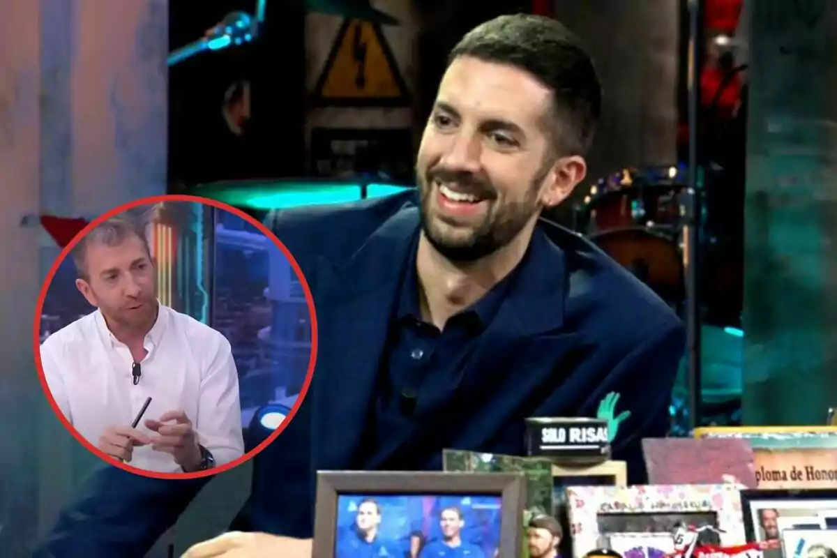 Un hombre sonriente con barba y traje oscuro está sentado en un estudio de televisión, en la esquina inferior izquierda hay un círculo rojo con la imagen de otro hombre con camisa blanca hablando.