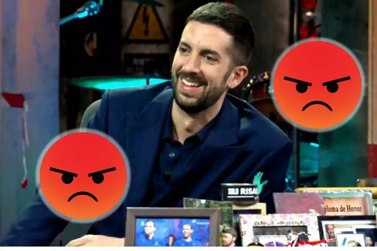 Un hombre sonriente en un set de televisión con dos emojis de enojo superpuestos.