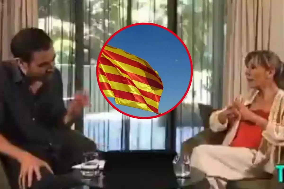 Dos personas conversan sentadas en una sala con cortinas claras y una ventana al fondo, mientras en el centro de la imagen aparece un círculo con la bandera catalana ondeando sobre un cielo azul