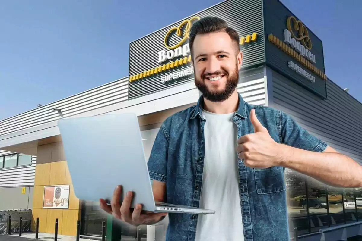 Un hombre sonriente con barba sostiene una laptop y hace un gesto de aprobación con el pulgar frente a un supermercado Bonpreu.