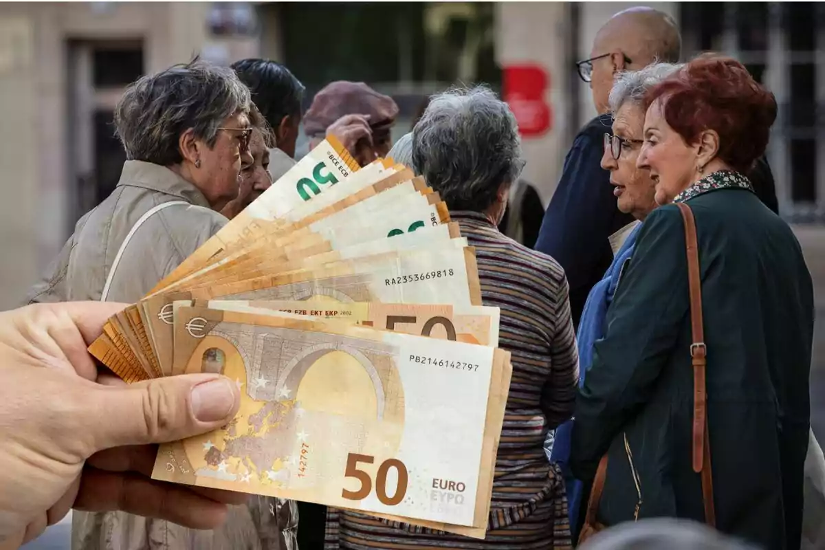 Un grupo de personas mayores conversando mientras una mano sostiene billetes de euro en primer plano.