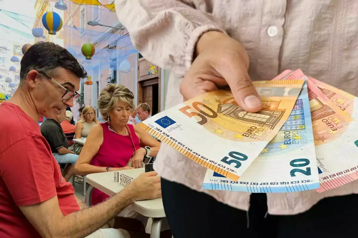 Una persona sostiene billetes de euro mientras un grupo de personas juega al dominó en una terraza decorada con globos de colores.