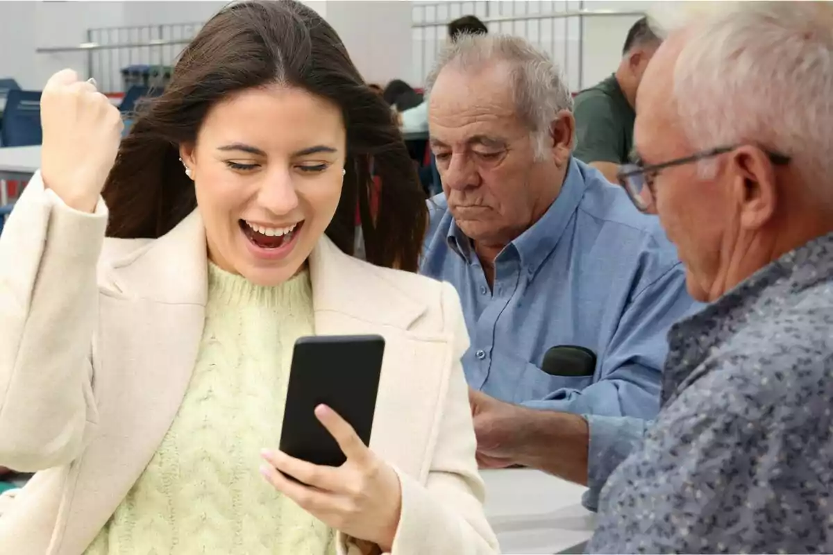 Una dona jove somriu emocionada mentre mira el telèfon mòbil, amb dos homes grans asseguts darrere seu.