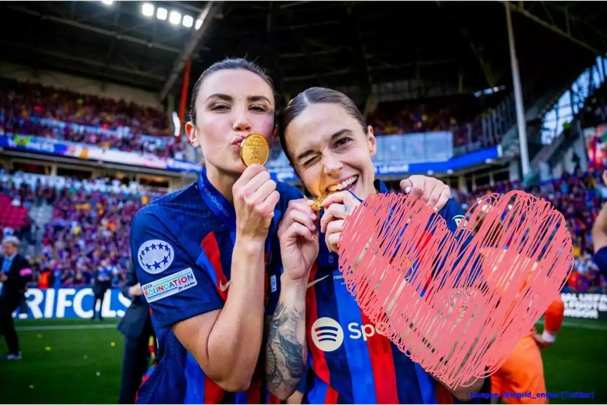 Dos futbolistes celebren amb medalles d'or en un estadi ple d'espectadors, una té un tatuatge visible al braç i hi ha un cor dibuixat sobre la imatge.