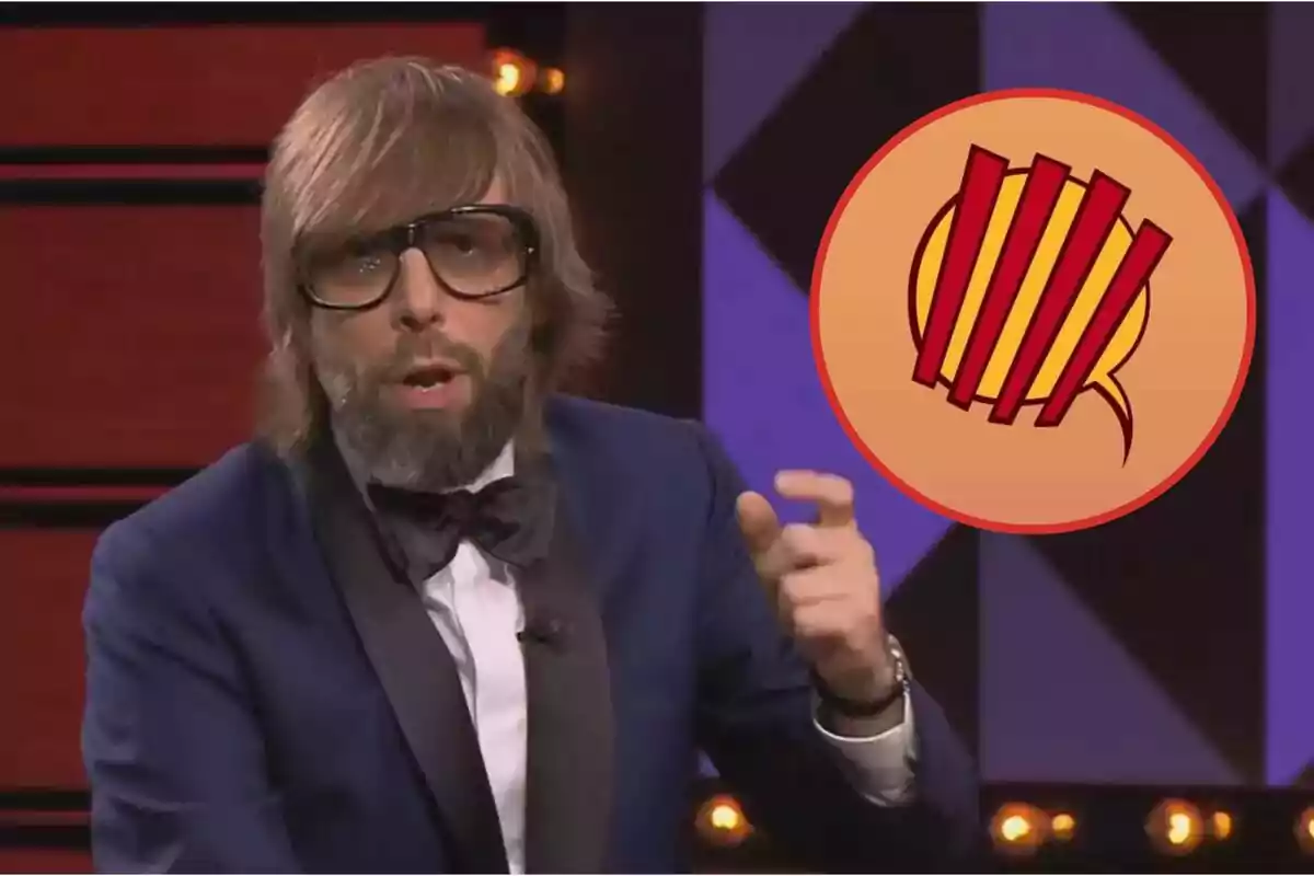 Un hombre con barba y gafas grandes lleva un esmoquin azul y corbata de moño mientras gesticula con la mano; a su lado hay un logotipo circular con líneas rojas sobre un fondo amarillo.