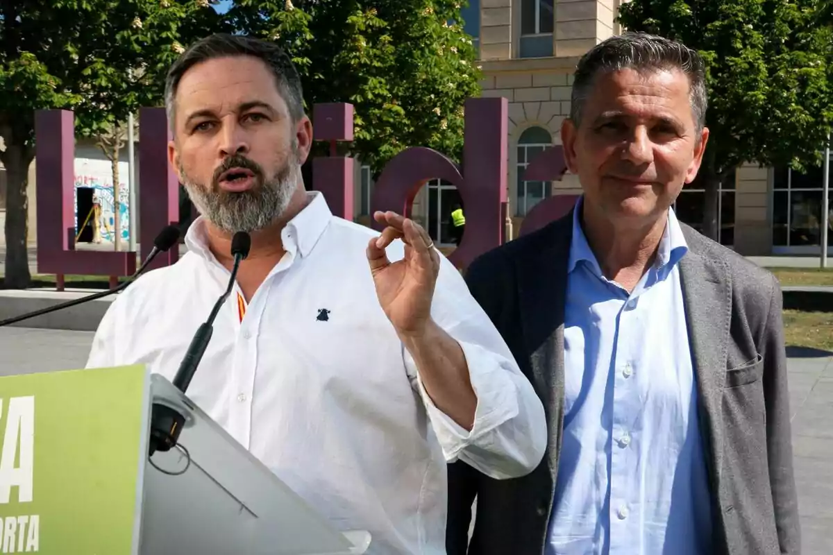 Dos hombres de pie al aire libre, uno hablando en un micrófono y el otro sonriendo, con un edificio y árboles de fondo.