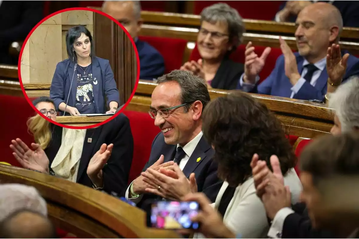 Una mujer hablando en un podio mientras varias personas aplauden en un parlamento.