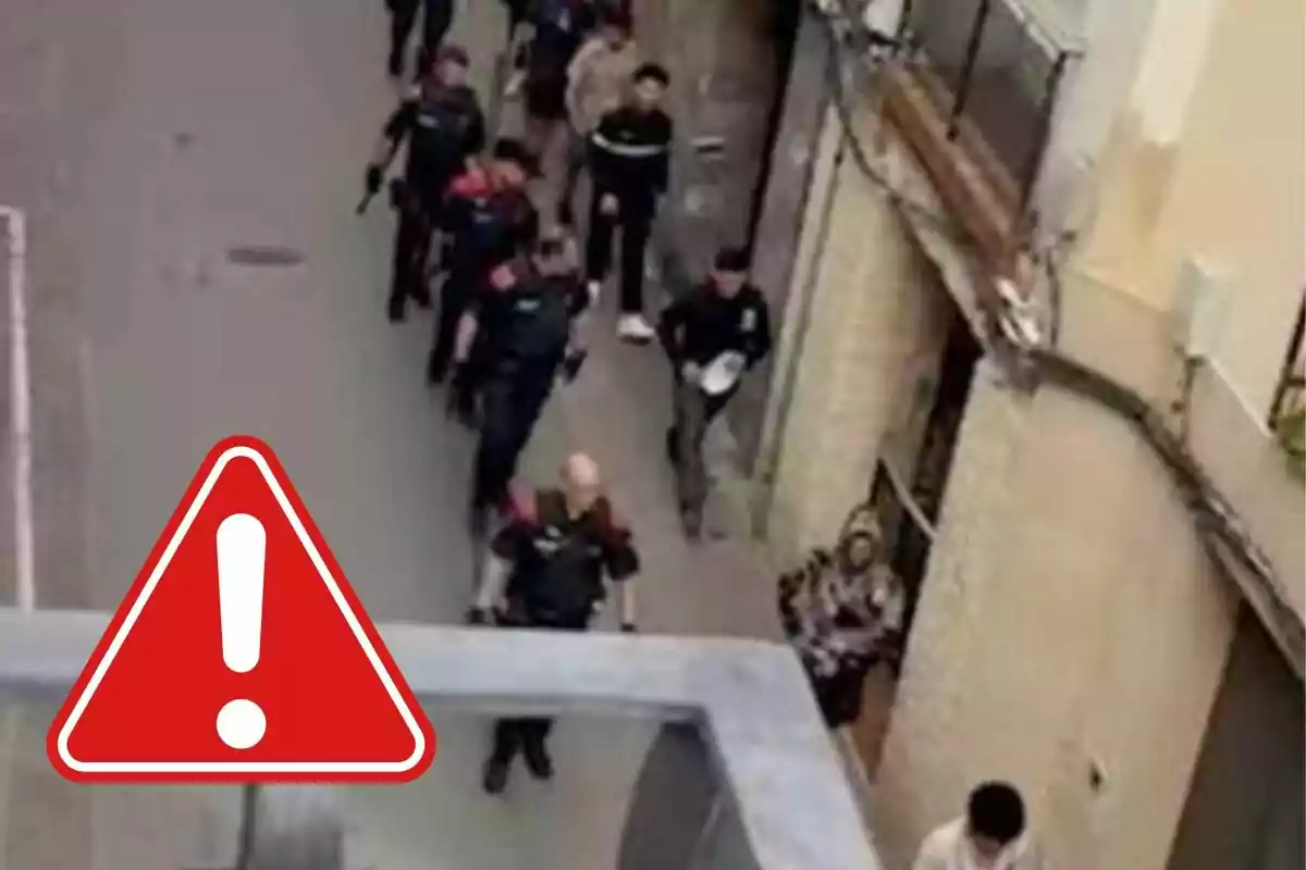 Un grupo de personas camina por una calle estrecha, algunas de ellas parecen ser oficiales de policía, y hay un símbolo de advertencia en la imagen.