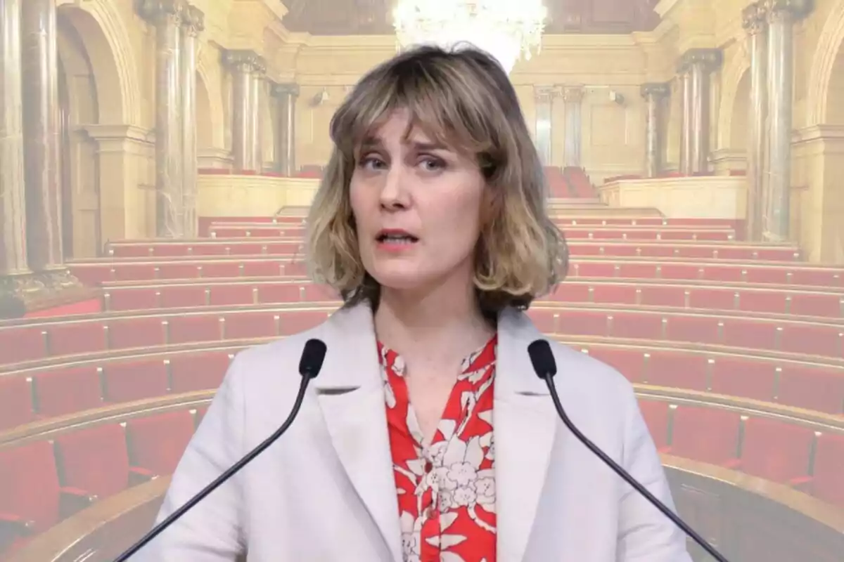 Una mujer hablando frente a dos micrófonos en un salón con asientos rojos y columnas decorativas.