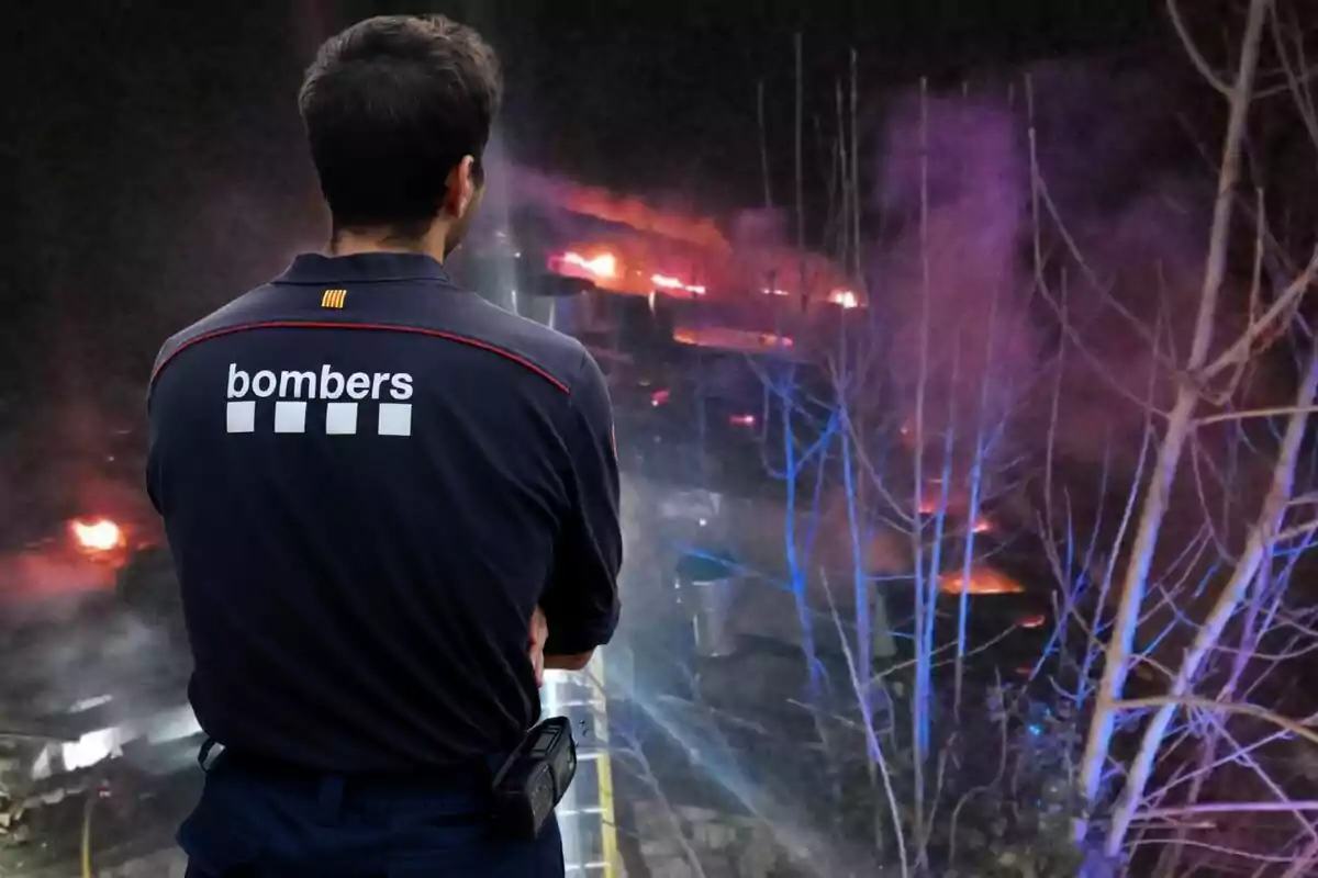 Un bomber d'esquena observa un incendi nocturn amb flames i fum al fons.