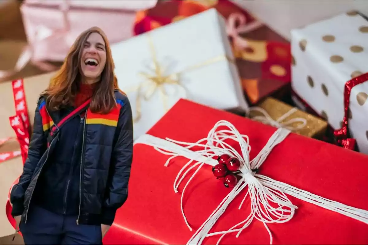 Una persona sonriente con chaqueta colorida está frente a una pila de regalos envueltos en papel decorativo.