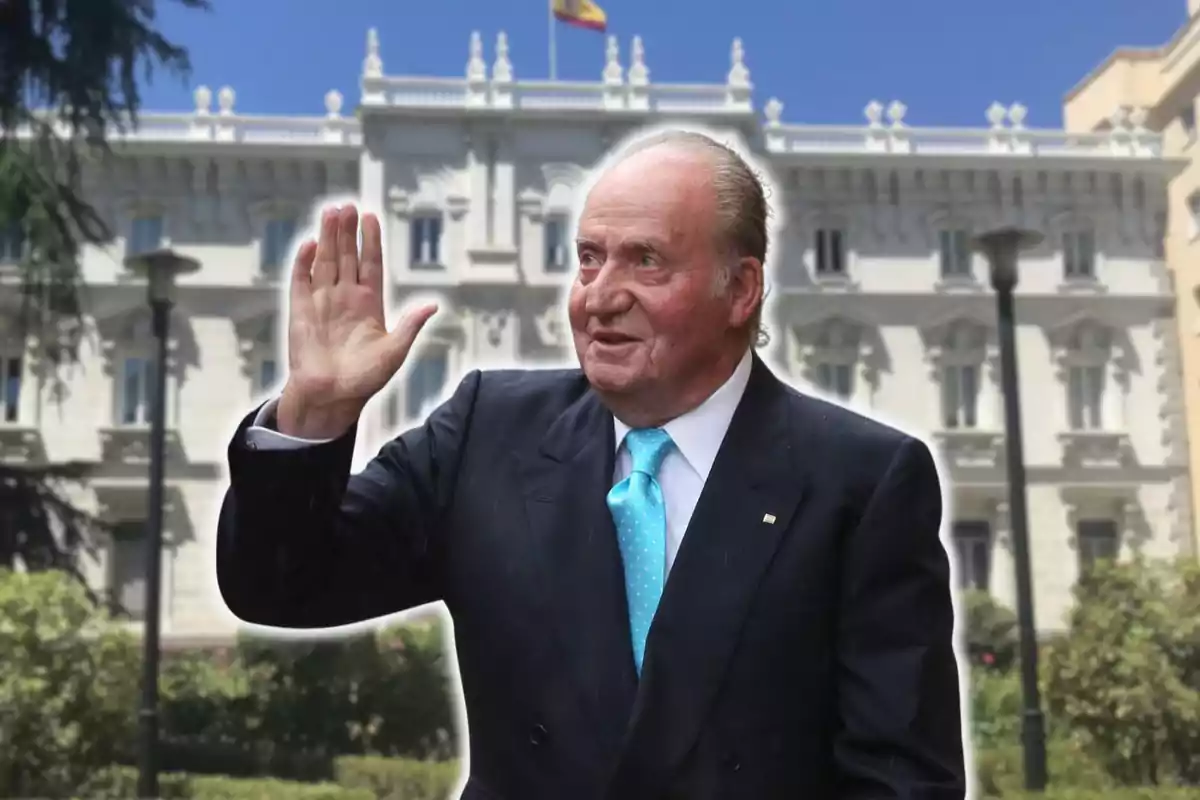 Un hombre de traje oscuro y corbata azul levanta la mano frente a un edificio blanco con detalles arquitectónicos y una bandera en la parte superior.