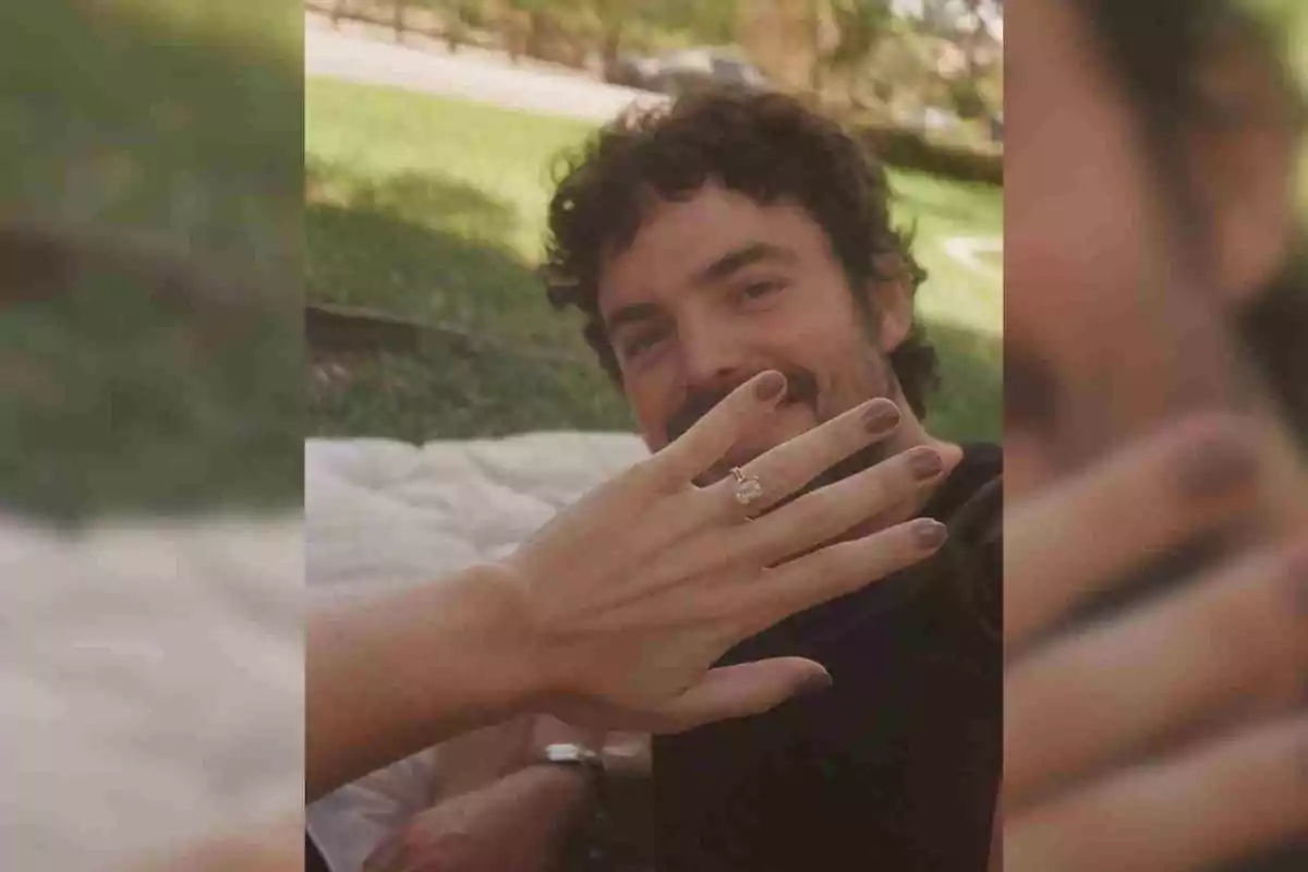 La mà d'Stella del Carmen amb un anell de compromís en primer pla i una persona somrient al fons en un parc. La mà d'Stella del Carmen amb un anell de compromís en primer pla i una persona somrient al fons en un parc.