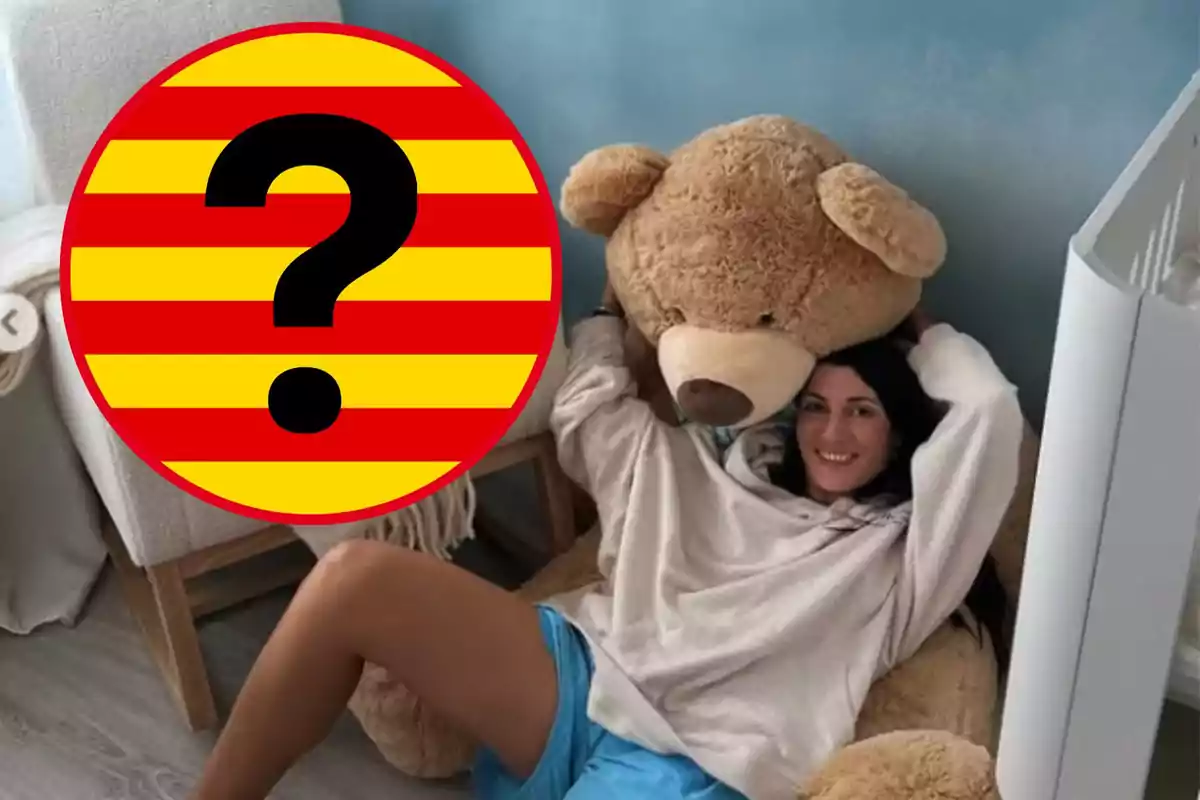 Una persona sonríe mientras está sentada en el suelo abrazando un gran oso de peluche en una habitación.