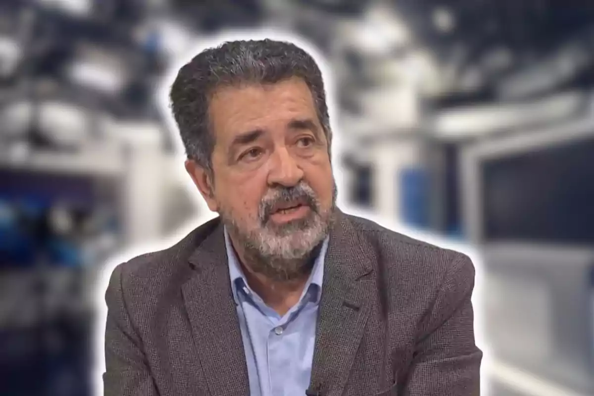 Un home amb barba i cabell canós, vestint una jaqueta grisa i camisa blava, en un entorn d'estudi de televisió.