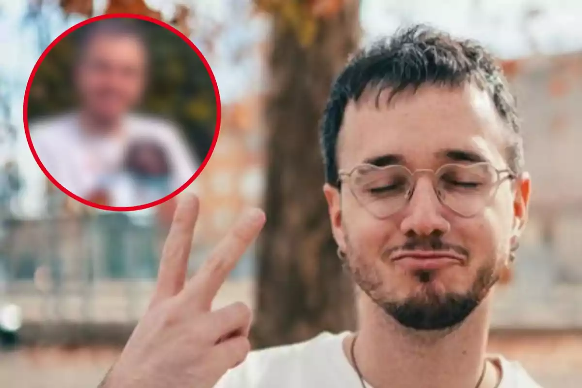 Un hombre con gafas y barba hace un gesto de paz con los dedos mientras cierra los ojos, con un fondo desenfocado de árboles y edificios.