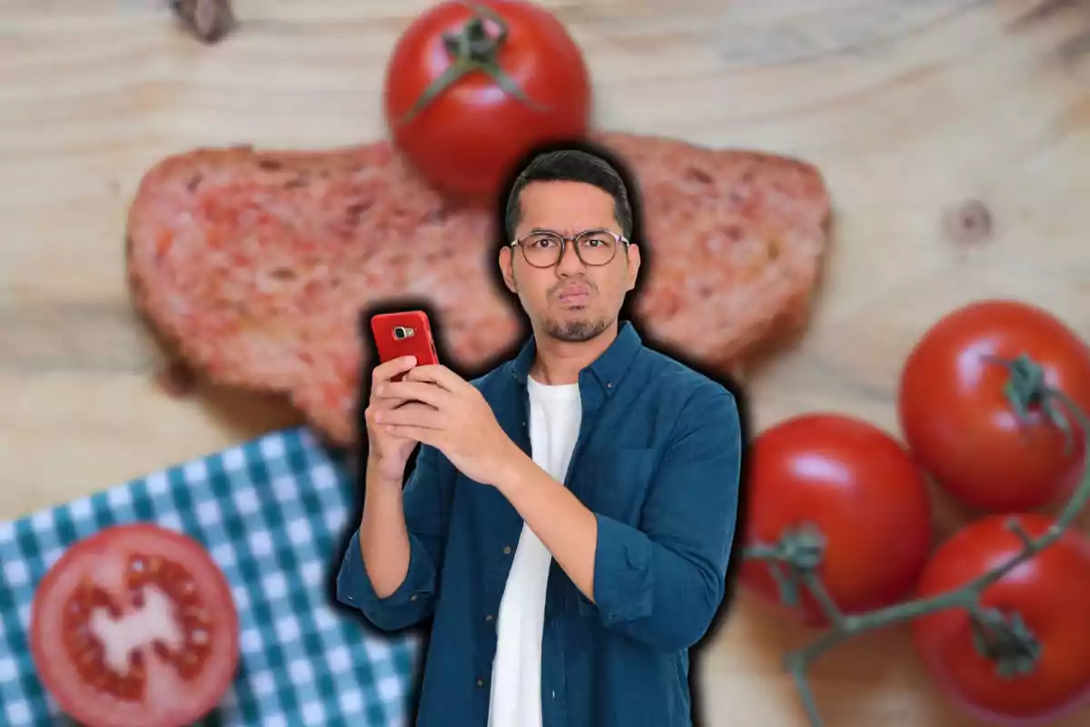 Hombre con expresión confundida sosteniendo un teléfono rojo frente a una rebanada de pan y tomates sobre una mesa de madera.