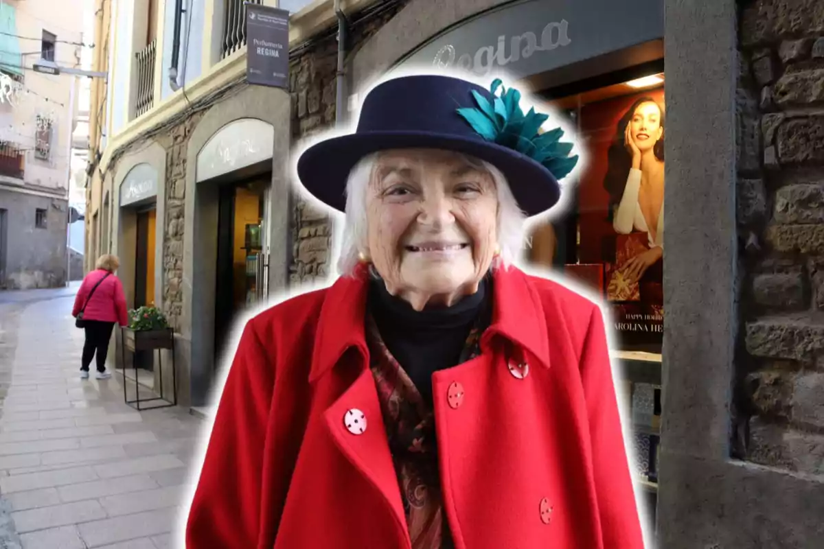 Una dona gran somriu mentre porta un abric vermell i un barret blau amb una flor, dreta davant d'una perfumeria en un carrer estret.