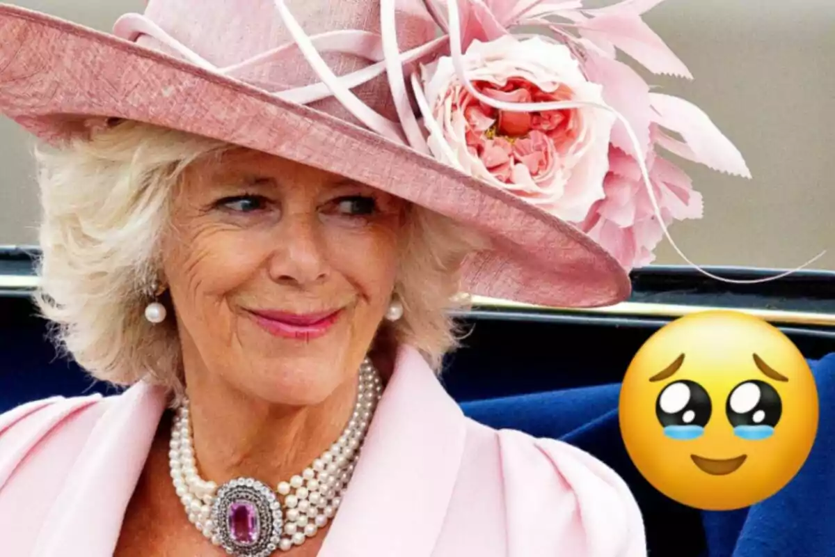 La reina Camila amb un elegant barret rosa adornat amb flors i un collaret de perles somriu mentre està asseguda en un vehicle, acompanyada d'un emoji amb ulls brillants i llàgrimes d'emoció.