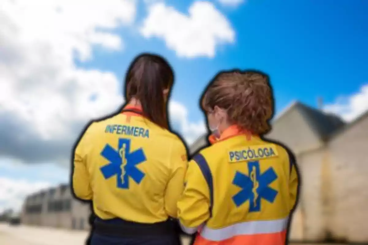 Dos professionals de la salut amb uniformes grocs d'emergència, una infermera i una psicòloga, d'esquena en un entorn exterior amb cel blau i núvols.