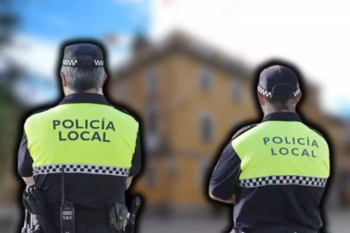 Dos agentes de policía local de espaldas con chalecos reflectantes amarillos y fondo desenfocado.