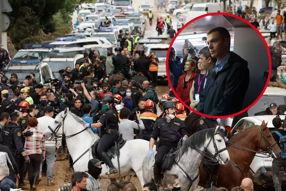 Una multitud de personas y fuerzas de seguridad se congregan en una calle llena de vehículos de emergencia, mientras un grupo de personas observa desde un lugar cerrado.