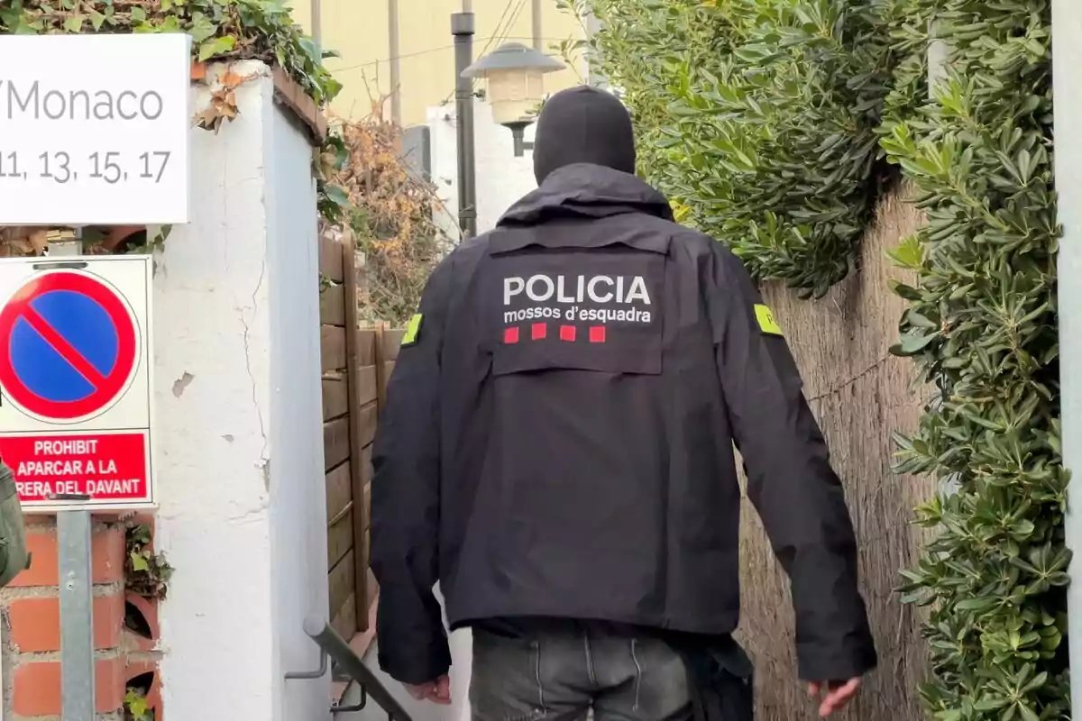 Un agent de la policia dels Mossos d'Esquadra camina per un passadís exterior envoltat de vegetació, amb un cartell que indica la prohibició d'aparcar.