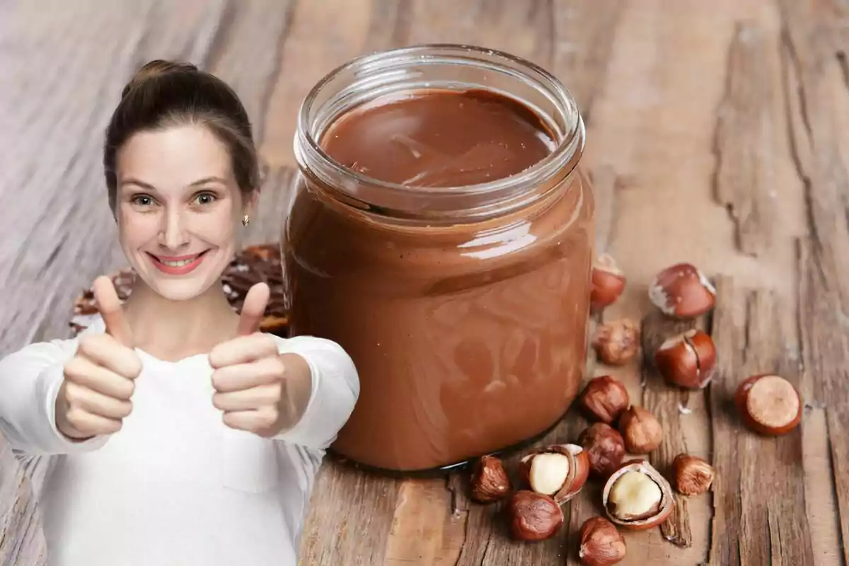 Una persona sonriente con pulgares arriba junto a un frasco de crema de avellanas sobre una mesa de madera.