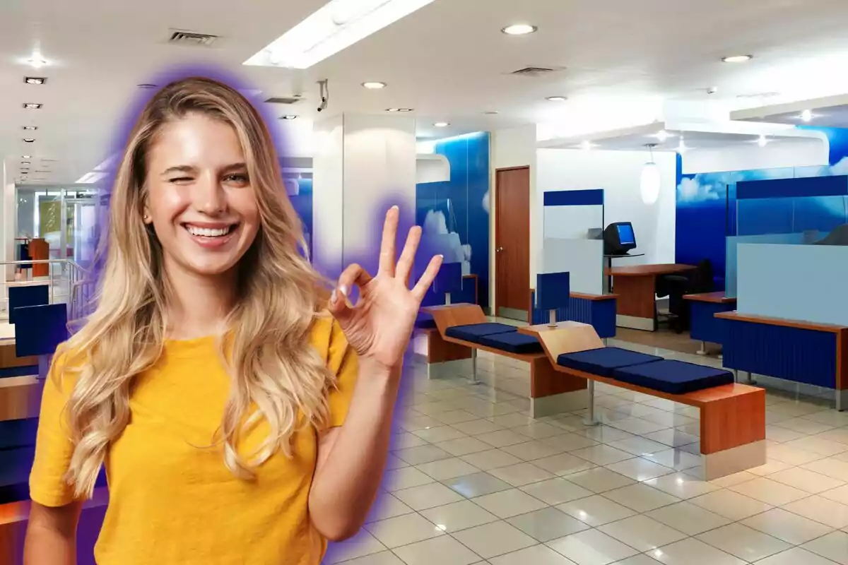 Una dona somrient amb samarreta groga fa un gest d'ok amb la mà en un entorn modern d'oficina o banc amb mobles blaus i de fusta. Una dona somrient amb samarreta groga fa un gest d'ok amb la mà en un entorn modern d'oficina o banc amb mobles blaus i de fusta.