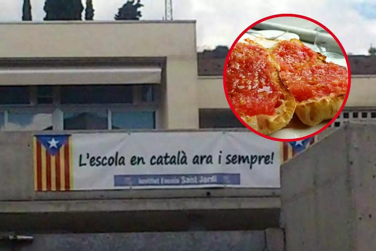 Un cartell en un edifici mostra la frase "L'escola en català ara i sempre" al costat d'una bandera catalana, ia la cantonada superior dreta hi ha una imatge de pa amb tomàquet.