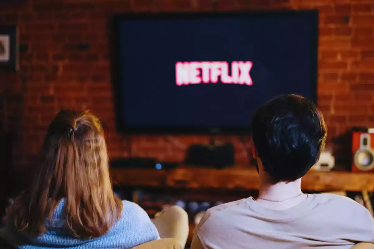 Dos personas sentadas en un sofá viendo una pantalla de televisión con el logotipo de Netflix en un ambiente acogedor con paredes de ladrillo.