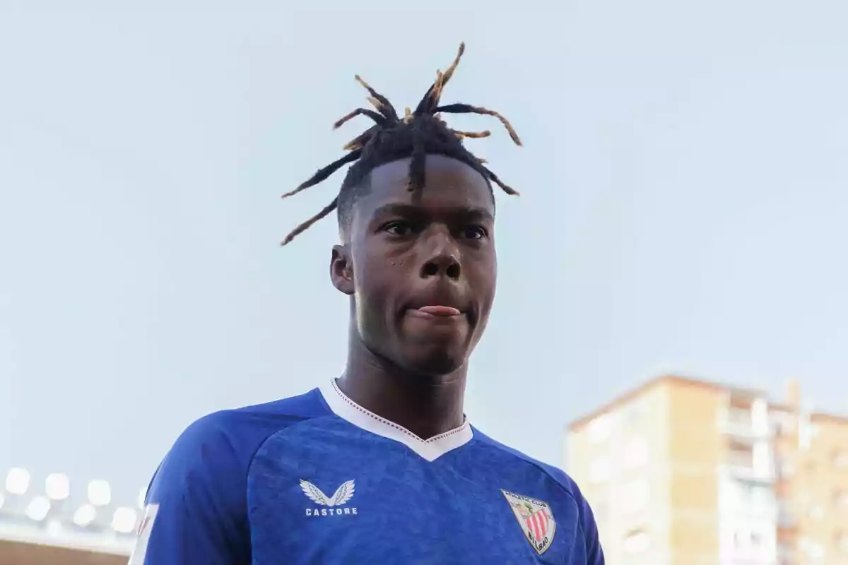 Un jugador de futbol amb samarreta blava i el logo de Castore, amb els cabells recollits en rastes, treu la llengua mentre mira cap endavant. Un jugador de futbol amb samarreta blava i el logo de Castore, amb els cabells recollits en rastes, treu la llengua mentre mira cap endavant.